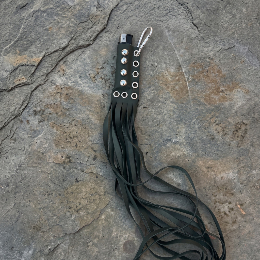 A black leather flogger keychain with silver-tone hardware, including a lobster clasp, resting on a textured gray stone surface.