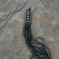 A black leather flogger keychain with silver-tone hardware, including a lobster clasp, resting on a textured gray stone surface.